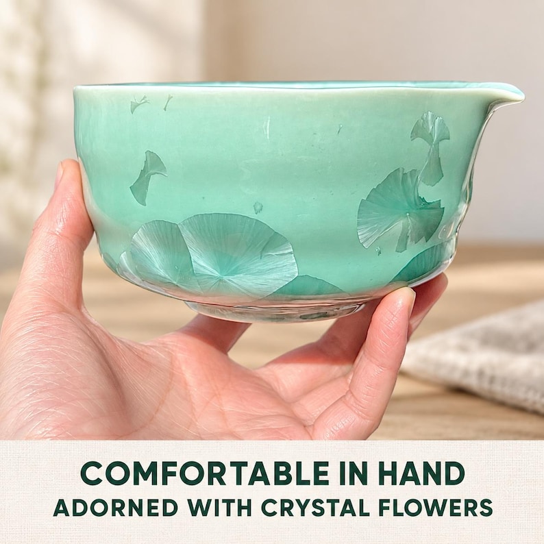 May include: A light mint green ceramic bowl featuring a crystalline glaze pattern that resembles delicate flower shapes. The bowl has a smooth, rounded body with a small pouring spout on the rim. Text on the image reads: COMFORTABLE IN HAND ADORNED WITH CRYSTAL FLOWERS.