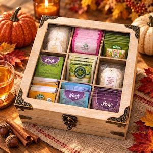 May include: A square wooden tea storage box with a clear lid and nine internal compartments. The box features decorative metal corner accents and a front latch. Inside are various tea bags from brands including Yogi, Organic India, and Tadin.