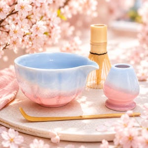 Op de afbeelding: Een matcha theeset met een blauw-roze gradiënt kom, een bamboe klopper, een kleine bijpassende container en een gouden lepel. De set is gerangschikt op een licht oppervlak, met kersenbloesems op de achtergrond.
