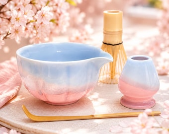 May include: A ceramic matcha bowl with a light blue and pink gradient finish, a matching ceramic whisk holder, and a bamboo tea scoop. The bowl features a scalloped rim and a small pouring spout, while the whisk holder has a smooth, rounded shape.