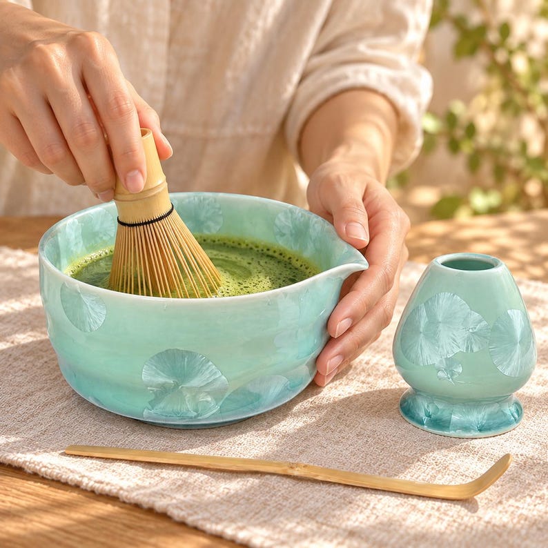 May include: A light green ceramic matcha bowl with a floral pattern, a bamboo whisk submerged in green tea, a matching ceramic whisk holder, and a long, thin bamboo tea scoop. The set features a glossy finish with subtle, crystalline petal designs.