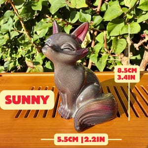Puede incluir: Una figura de zorro de cer&aacute;mica gris oscuro con orejas interiores de color rojo rosado y un acabado liso y brillante. El zorro est&aacute; sentado con los ojos cerrados. La figura mide 8,5 cm de alto y 5,5 cm de ancho, etiquetada como SUNNY.
