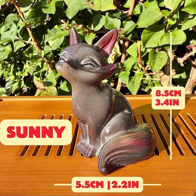 Puede incluir: Una figura de zorro de cer&aacute;mica gris oscuro con orejas interiores de color rojo rosado y un acabado liso y brillante. El zorro est&aacute; sentado con los ojos cerrados. La figura mide 8,5 cm de alto y 5,5 cm de ancho, etiquetada como SUNNY.