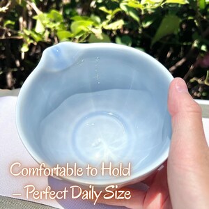 May include: A light blue ceramic bowl with a smooth, glossy finish and a small pouring spout on the rim. The interior of the bowl is rounded and deep, featuring a pale blue hue that reflects light. The text Comfortable to Hold &ndash; Perfect Daily Size is visible.