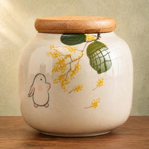 May include: A cream-colored ceramic canister with a rounded body and a cylindrical wooden lid. The canister features a painted illustration of a small white rabbit with pink cheeks standing near a branch of yellow mimosa flowers and green leaves.