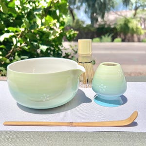 May include: A matcha tea set comprising a light green and blue ceramic bowl with a spout, a small vase, a bamboo whisk, and a bamboo scoop. The bowl and vase have white floral accents. The set is arranged on a light grey surface.