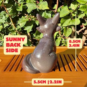Puede incluir: Una figura de zorro de cer&aacute;mica gris oscuro vista desde atr&aacute;s, con orejas puntiagudas y una cola curvada. La figura mide 8,5 cm de alto y 5,5 cm de ancho. El texto en la imagen dice SUNNY BACK SIDE.