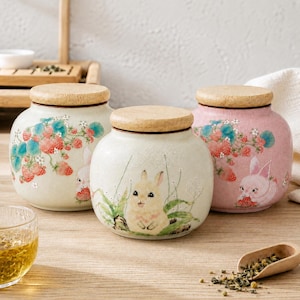May include: Three round ceramic storage jars with light wooden lids. The left jar is cream with strawberry vines and a rabbit. The center jar is cream with a rabbit in grass. The right jar is pink with strawberry vines and a rabbit.