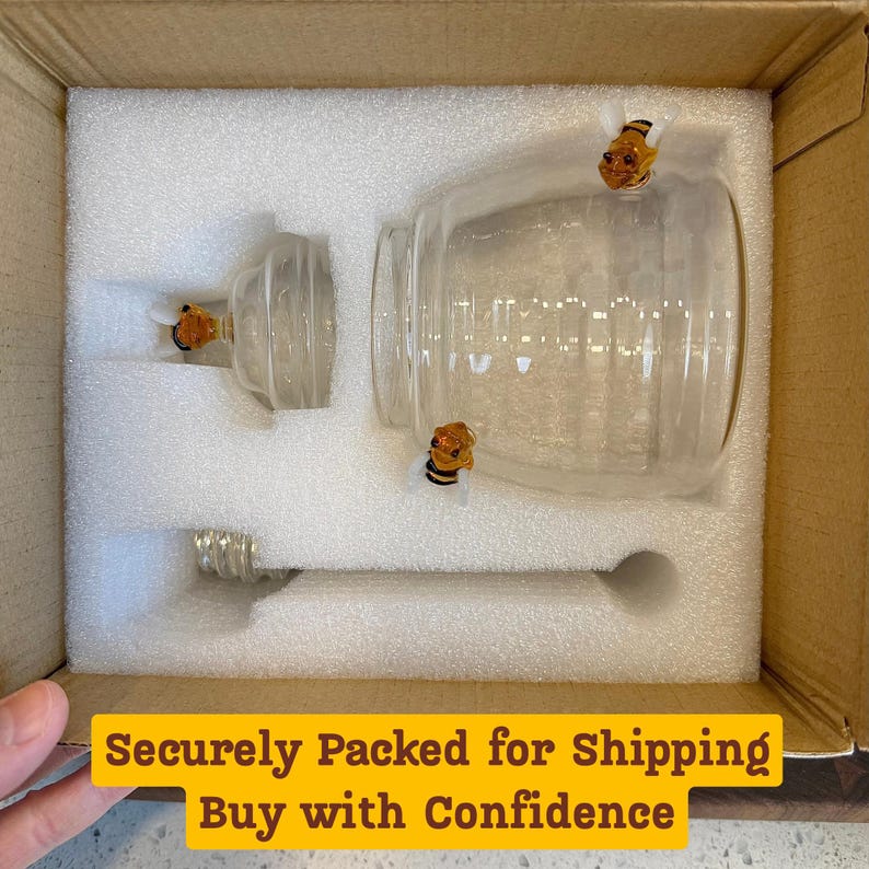 May include: A clear glass honey jar set with a lid, and a honey dipper, nestled in protective foam inside a cardboard box. The jar and lid are adorned with small, decorative bee accents. The set is designed for serving honey.