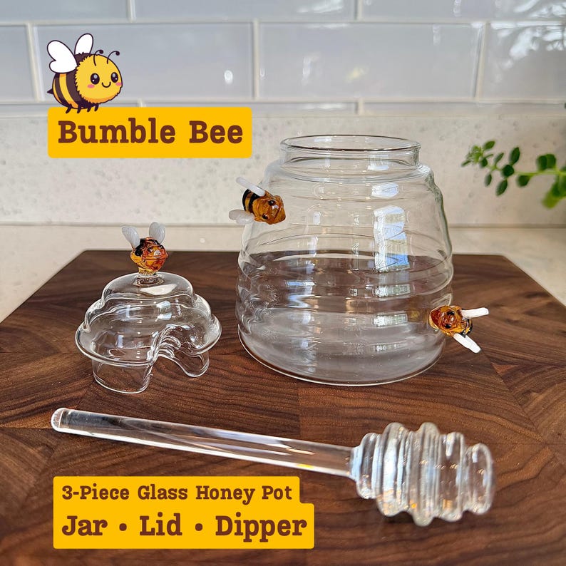 May include: A 3-piece glass honey pot set with a jar, lid, and dipper. The clear glass jar has a round shape and is adorned with small bumble bee figurines. The lid is shaped like a beehive. The set includes a glass honey dipper.