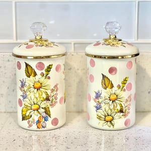 May include: Two white ceramic canisters with pink polka dots and floral designs. The canisters have gold rims and clear glass knobs on the lids.