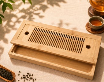 May include: A rectangular bamboo tea tray featuring a removable top panel with vertical drainage slats and two circular cutouts. The light-colored wooden tray has a hollow base designed to collect excess liquid during the tea brewing process.