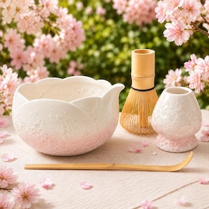 May include: A ceramic matcha tea set featuring a white and soft pink bowl with a scalloped petal rim, a bamboo whisk, a ceramic whisk holder with a matching floral texture, and a long, slender bamboo tea scoop for preparing traditional matcha.