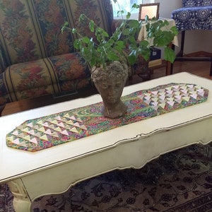 May include: A white coffee table with a decorative runner featuring a paisley pattern and pink and yellow origami triangles. A potted plant with green leaves sits on the table.