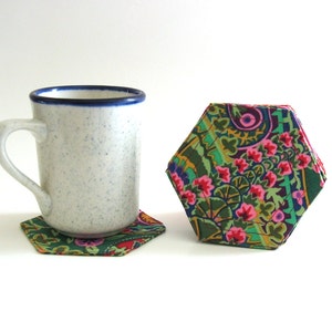 Pode incluir: Uma caneca de cerâmica branca com uma borda azul está sobre um descanso de copo hexagonal de tecido verde e rosa. À direita, está uma pilha de descansos de copo semelhantes.