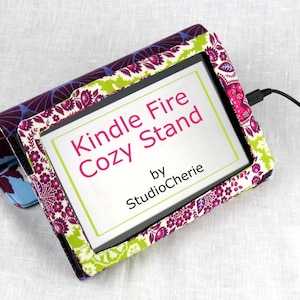 May include: A colorful fabric Kindle Fire Cozy Stand with a floral pattern. The stand has a cutout for the Kindle Fire and the text "Kindle Fire Cozy Stand by StudioCherie" is printed on the front.