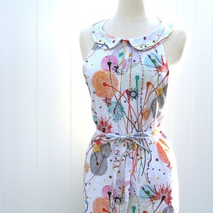 May include: A white sleeveless dress with a colorful abstract print of circles and lines. The dress has a white Peter Pan collar and a matching white sash tied at the waist.