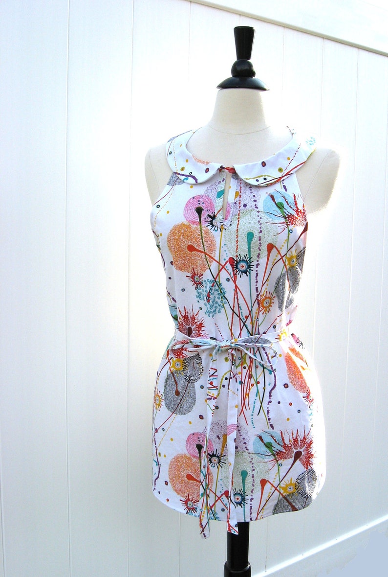 May include: A white sleeveless dress with a colorful abstract print of circles and lines. The dress has a white Peter Pan collar and a matching white sash tied at the waist.
