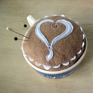 May include: A brown felt pincushion shaped like a teacup with a white heart design and white scalloped edges. The pincushion has three black pins sticking out of it.