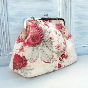 Puede incluir: Un bolso de mano vintage de estilo floral con cierre de plata y cadena. El bolso está hecho de tela de color crema con un estampado floral rojo y blanco.