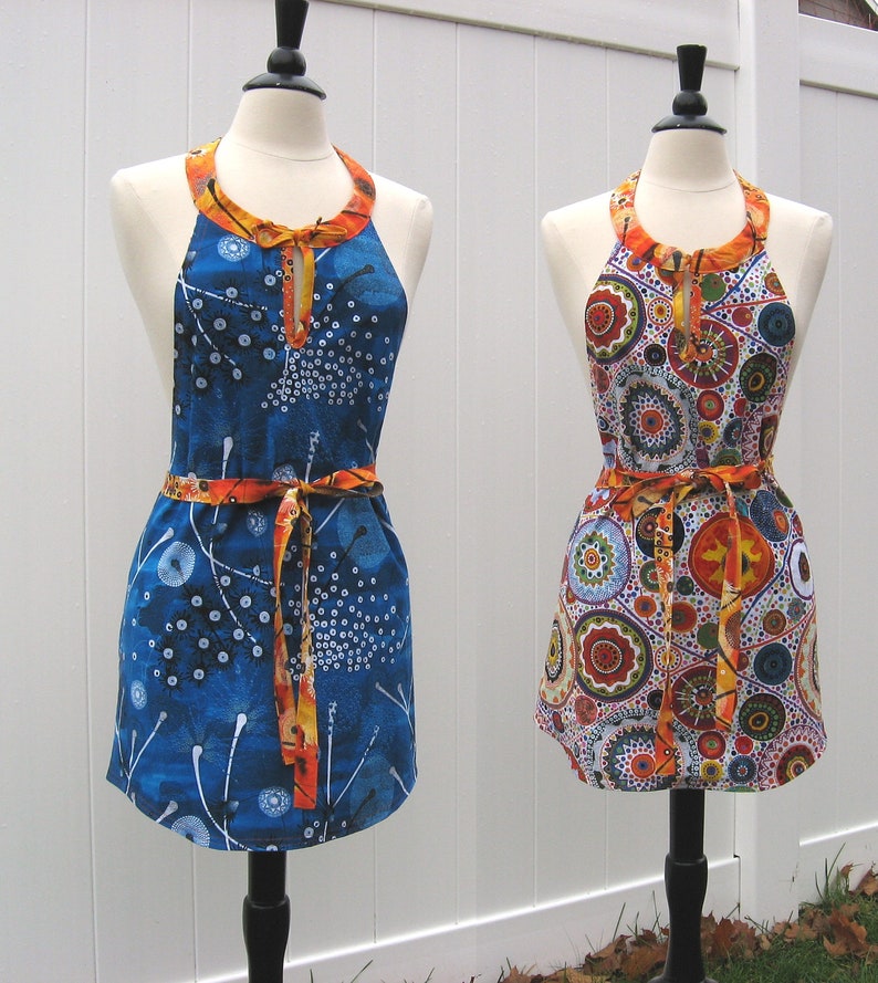 May include: Two colorful aprons with a halter neck style. One apron is blue with white and orange accents and the other is white with orange, red, and blue accents.
