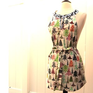 May include: A white apron with a black and white trim around the neck. The apron has a festive Christmas tree pattern with red, green, and blue trees on a white background.