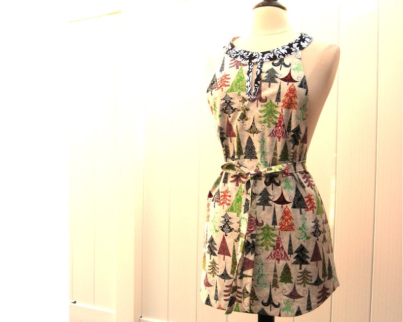 May include: A white apron with a black and white trim around the neck. The apron has a festive Christmas tree pattern with red, green, and blue trees on a white background.