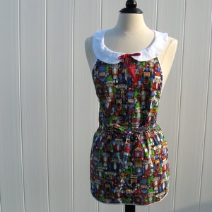 May include: A white collared apron with a red bow and a colorful print of nutcrackers. The apron has a gathered skirt and a tie at the waist.