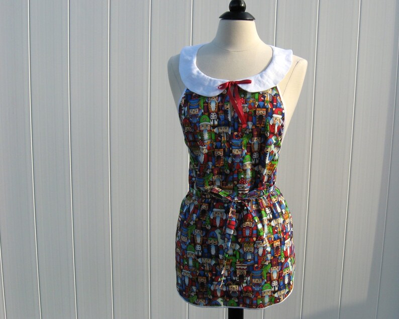 May include: A white collared apron with a red bow and a colorful print of nutcrackers. The apron has a gathered skirt and a tie at the waist.