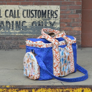 Op de afbeelding: Een blauwe en witte geprinte tote bag met een kleinere bijpassende tas eraan bevestigd. De tote bag heeft een ritssluiting en twee bovenste handvatten. De kleinere tas heeft een ritssluiting en één bovenste handvat. De tas ligt op een betonnen oppervlak voor een bakstenen muur.