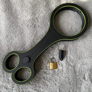 May include: Black and neon green restraint device with three circular openings and a large ring. Includes a small black cylinder, a gold padlock, and keys. The device is made of a textured material.