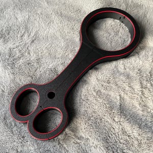 Puede incluir: Dispositivo de sujeción negro con ribete rojo. El dispositivo tiene dos puños circulares y un puño circular más grande conectado por una barra.