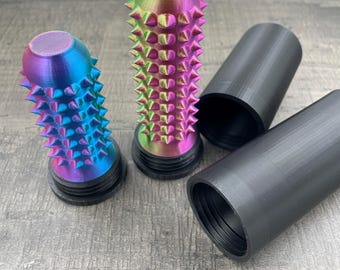 May include: Two spiked, iridescent bottle stoppers with black bases and two black cylindrical containers. The stoppers have a rainbow color gradient, with sharp, conical spikes covering their surfaces. The containers are smooth and matte black.
