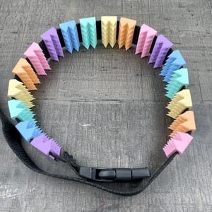Puede incluir: Un collar de masticación de silicona arcoíris con una correa negra ajustable y un cierre de hebilla. El collar está hecho de piezas individuales triangulares texturizadas.