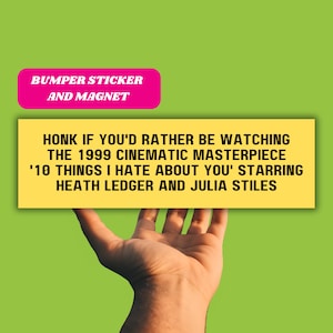 May include: Yellow bumper sticker and magnet with the text "HONK IF YOU'D RATHER BE WATCHING THE 1999 CINEMATIC MASTERPIECE '10 THINGS I HATE ABOUT YOU' STARRING HEATH LEDGER AND JULIA STILES". A hand is holding the sticker in front of a green background.