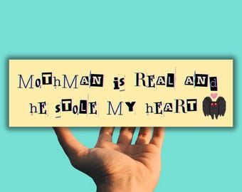 May include: A yellow sign with the text "MothMan is REAL and he STOLE MY HEART" in black letters with a black moth with red eyes and pink heart above the text.