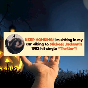 Könnte beinhalten: Ein gelber und orangefarbener Stoßfängeraufkleber mit einem Waschbären, der einen Hexenhut trägt und einen Besen hält. Der Text auf dem Aufkleber lautet "KEEP HONKING! I'm sitting in my car vibing to Michael Jackson's 1982 hit single "Thriller"!"