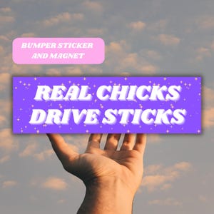 Puede incluir: Pegatina de parachoques morada con texto blanco que dice "REAL CHICKS DRIVE STICKS" con estrellas de purpurina rosa sobre un fondo blanco. La pegatina se sostiene en una mano contra un cielo nublado.