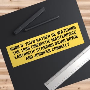 May include: Yellow bumper sticker with black text that reads "HONK IF YOU'D RATHER BE WATCHING THE 1986 CINEMATIC MASTERPIECE 'LABYRINTH' STARRING DAVID BOWIE AND JENNIFER CONNELLY"