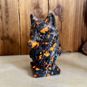 以下が含まれることがあります： 黒いベースとオレンジ色のアクセントが施された、石で彫られたオオカミの彫刻。オオカミは直立しており、耳を立てています。彫刻は明るい色の表面に置かれ、背景は木です。
