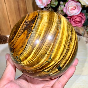 Könnte beinhalten: Eine polierte Tigeraugenkugel, die Bänder in goldgelben und braunen Farbtönen zeigt. Der Edelstein wird in einer Hand gehalten, mit einem verschwommenen Hintergrund aus Blumen und Holz. Diese dekorative Kugel ist ein Naturstein.