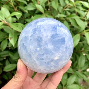 Pode incluir: Uma esfera de cristal azul claro com aparência nublada. A esfera é segurada em uma mão, com um fundo de folhas verdes. A bola de cristal é um item decorativo.