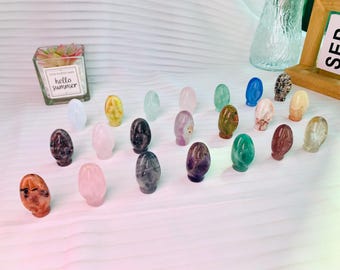 May include: A collection of 20 small, polished, egg-shaped crystal stones in various colors, including green, purple, pink, black, white, and brown. The stones are arranged on a white surface.