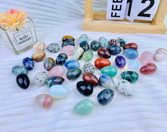 May include: A collection of 30 tumbled gemstone eggs in various colors and patterns, including green, blue, red, pink, and brown. The eggs are arranged on a white surface.