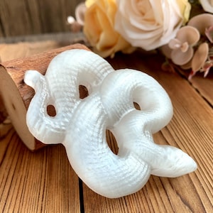 May include: A white, iridescent, ceramic snake sculpture. The snake is curled up in a coil, with its head resting on its body. The sculpture is made of a smooth, shiny material.