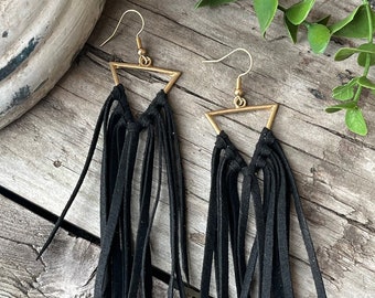 May include: A pair of black suede fringe earrings with gold triangle frames. The earrings are hanging from a wooden surface.