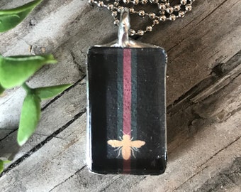 May include: A silver chain necklace with a rectangular pendant featuring a black and green striped pattern with a red stripe and a gold bee graphic.