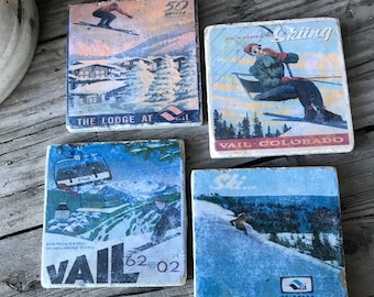 May include: Four square ceramic coasters with images of skiers and the Vail, Colorado ski resort. The coasters feature a blue and white ski scene, a red and white ski scene, a blue and white ski lift scene, and a red and white ski scene with the text "The Lodge at Vail".