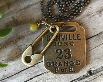 May include: A brass necklace with a charm that says "Danville June 23 Grange P. of H.". The charm is attached to a safety pin and a green bead.