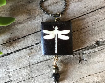 May include: A black square pendant with a white dragonfly design hangs from a brown chain necklace. The pendant has a small black and gold bead dangle.